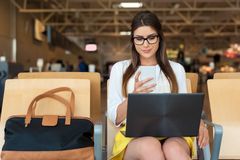 En kvinde venter i lufthavnen og kigger på sin smartphone og laptop.