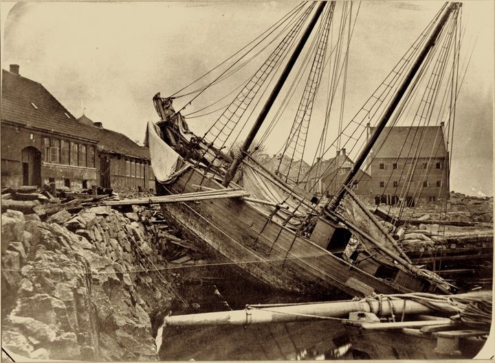 En af de værste stormfloder oplevede bornholmerne i november 1872. Dagen efter tog Rønnes fotograf, Gottlieb Støckel til Allinge og forevigede det frygtelige syn, der mødte ham. Her er han på den sønderslåede havn og ser hen mod Allinge Mølle, der har mistet vingerne. Skonnerten Robert af Rønne er blevet kastet op på kajen. Den borede bovsprydet ind gennem muren til den bagvedliggende købmandsgård. Robert blev senere bjærget og bragt til Rønne for at blive repareret. Foto: Bornholmske fotografer