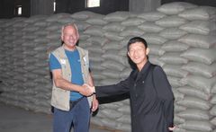Mission Østs generalsekretær Kim Hartzner hilser på en repræsentant fra de nordkoreanske myndigheder på et nødhjælpslager med bjerge af sække med majs og sojabønner fra Mission Øst. Fødevarerne deles ud til tusindvis af underernærede børn på nordkoreanske børneinstitutioner.