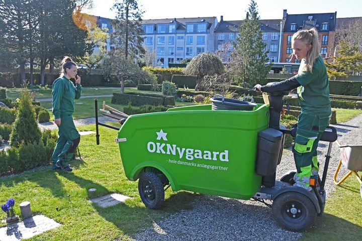 På Brønshøj Kirkegård kører Sofie Kruse og Freja Busch fra OKNygaards driftsafdeling i Glostrup rundt på en af virksomhedens el-ladvogne. El-ladvognene bliver især brugt på kirkegårde og hospitaler, hvor der er begrænset plads og behov for ro.