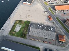 Luftfoto af et område ved vandet med asfalt, bygninger og grønne arealer.