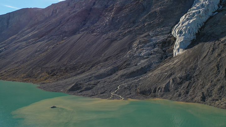 Grumset smeltevand flyder ud i Arktiske Hav, hvor det kan forhindre solens lys i at nå ned til havbunden. I vandet ses en 10 m lang båd med forskere, der tager vandprøver. Billedet er taget med drone i en fjord i nordøstlige Grønland.