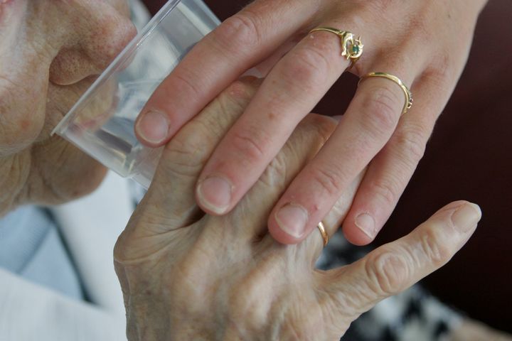 To hænder holdes om en plastikbæger, den ene hånd ældre med rynker, den anden yngre med smykker.