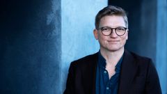 Billedet viser en mand med briller, der står foran en blågrå baggrund.