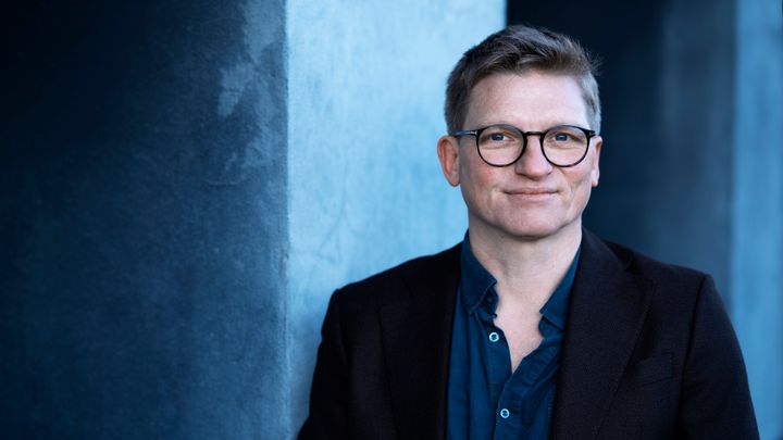 Billedet viser en mand med briller, der står foran en blågrå baggrund.