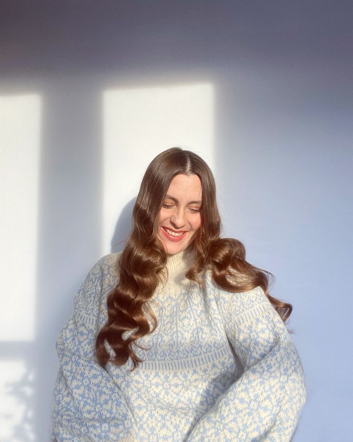 Billedet viser en smilende person i en mønstret sweater med langt bølget hår, der står i sollys.
