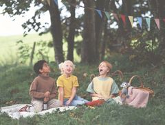 Tre børn sidder på et tæppe udendørs med et picnickurv, omgivet af natur og farverige flag.