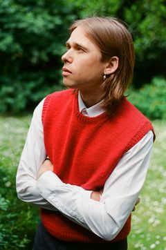 Ung mand med skulderlangt hår, iført rød sweater over en hvid skjorte, står med korslagte arme i en grøn have.
