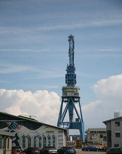 En stor blå kran ved enden af Balticagade i Aarhus, med bygninger og lager af genbrugsmaterialer i nærheden.