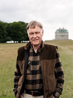 En mand i udendørs tøj står på en mark med træer og en bygning i baggrunden.