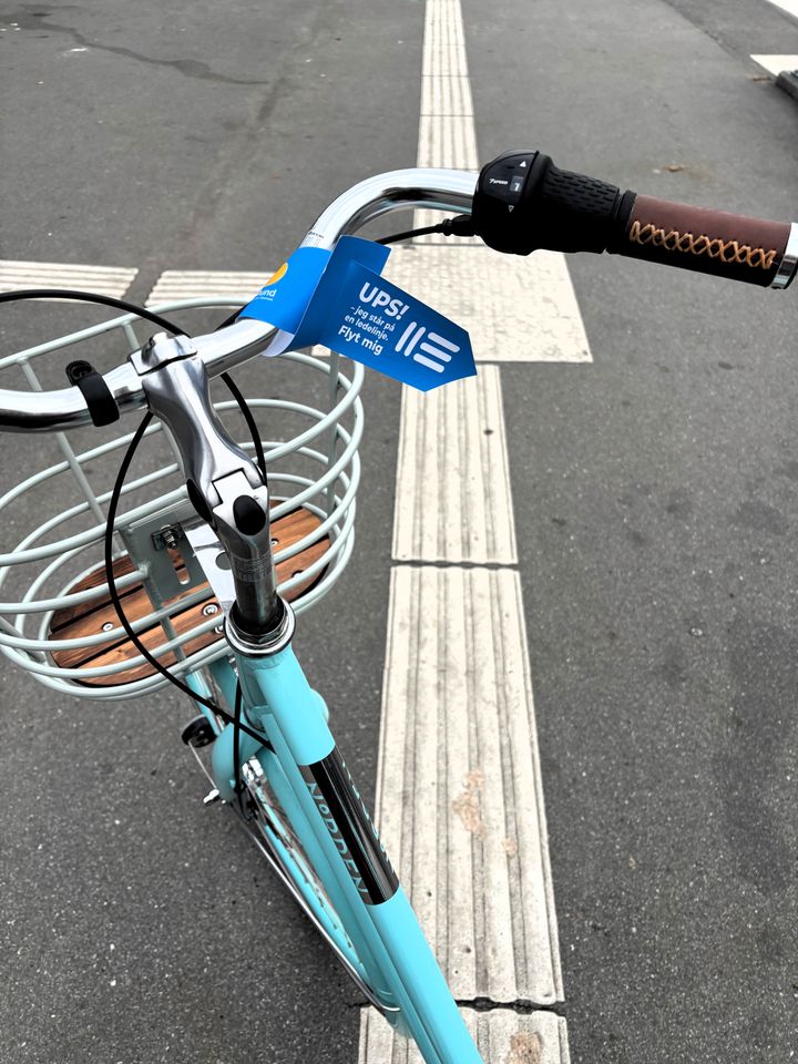 Blå cykel som spærrer ledelinje. På styret er der påsat et blåt mærkat, hvor der står: Ups! Jeg står på en ledelinje. Flyt mig