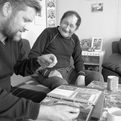 Christian Vium (til venstre i billedet) sidder i en sofa sammen med en beboer fra Ikerasak og kigger i et fotoalbum. Han har sammen med beboerne i Ikerasak anvendt private fotos og historisk materiale sammen med nye optagelser til udstillingen.