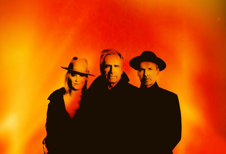 Tre personer kigger ud på beskueren. Det er Michael falch flankeret af to musikere. En dame til højre og en mand til venstre. Begge musikere har hat på. baggrunden er orange som flammer.