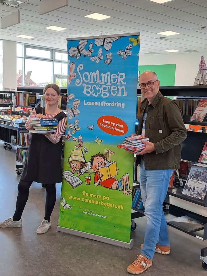 To mennesker står ved siden af en elevudfordringsplakat fra Sommerbogen i et bibliotek. De holder en stak bøger.