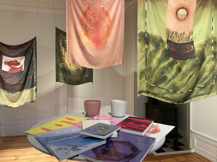 Et bord med bøger og kunstværker hængende i et rum, der kombinerer kunstudstilling og lyrik.