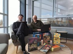 To personer sidder på sofaer med laptops, omgivet af bøger på et bord foran dem.