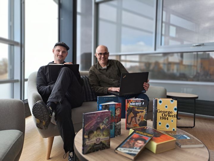 To personer sidder på sofaer med laptops, omgivet af bøger på et bord foran dem.