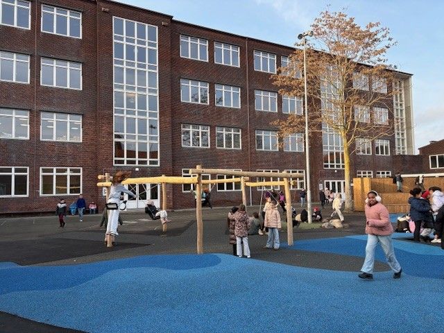 Børn leger på en skolegård med klatrestativ og blåt underlag foran en stor bygning.