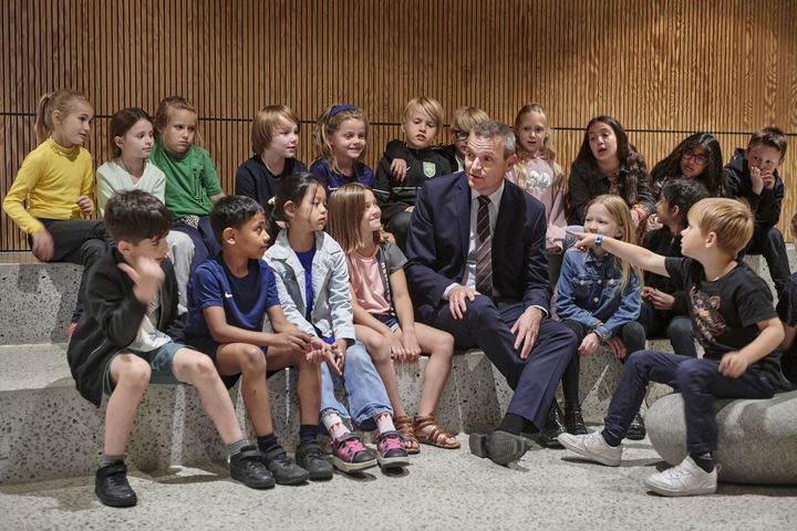 En gruppe børn sidder omkring en voksen mand i jakkesæt under et skolebesøg.