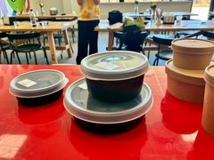 Flere plastikbeholdere med låg står på et rødt bord i et rum med stole og borde i baggrunden. I baggrunden står engangsboksene.