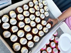 En kasse med mange cupcakes med hvid glasur og printede toppe, hvor der står "Skolen ved Himmelbjerget". En hånd tager en cupcake.