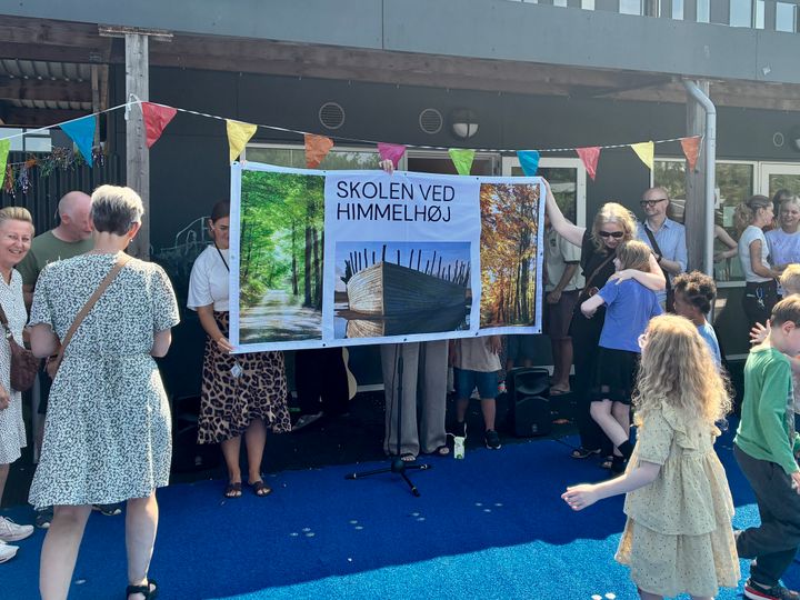 En gruppe mennesker foran et banner med teksten "Skolen ved Himmelbøj." Farverige flag hænger over dem.