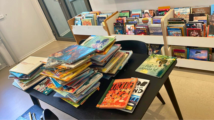 En skolebibliotek med mange børnebøger stablet på et bord og i hylder i baggrunden.