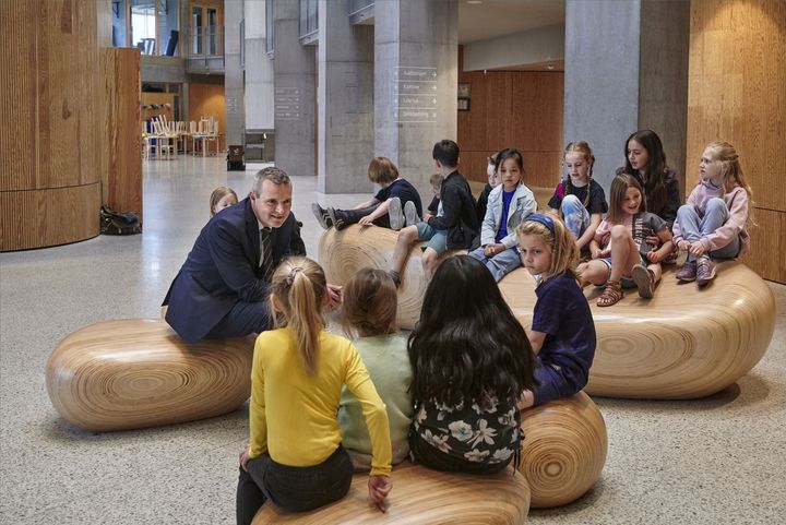 Borgmester taler med en gruppe børn, der sidder på moderne, organiske træmøbler i en skolebygning.