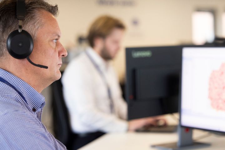 To mænd arbejder på computere på et kontor. En af dem har et headset på.