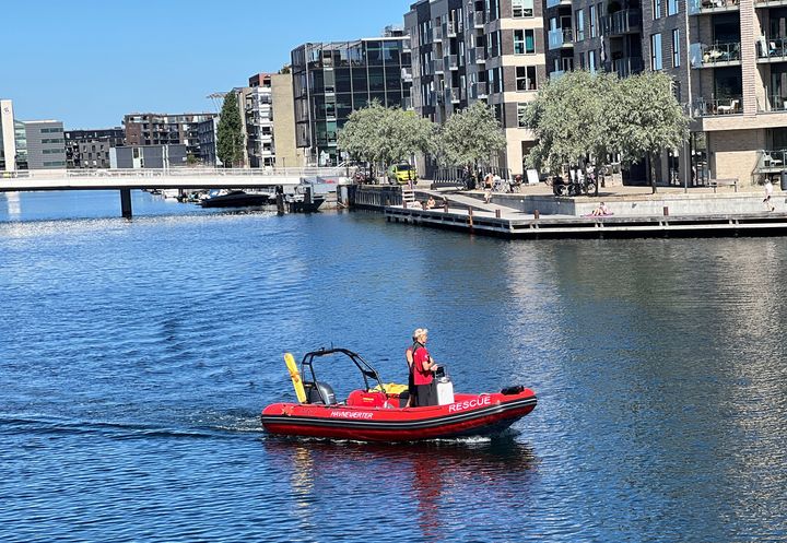 En redningsbåd sejler i Københavns havn med en person ombord, omgivet af moderne bygninger.