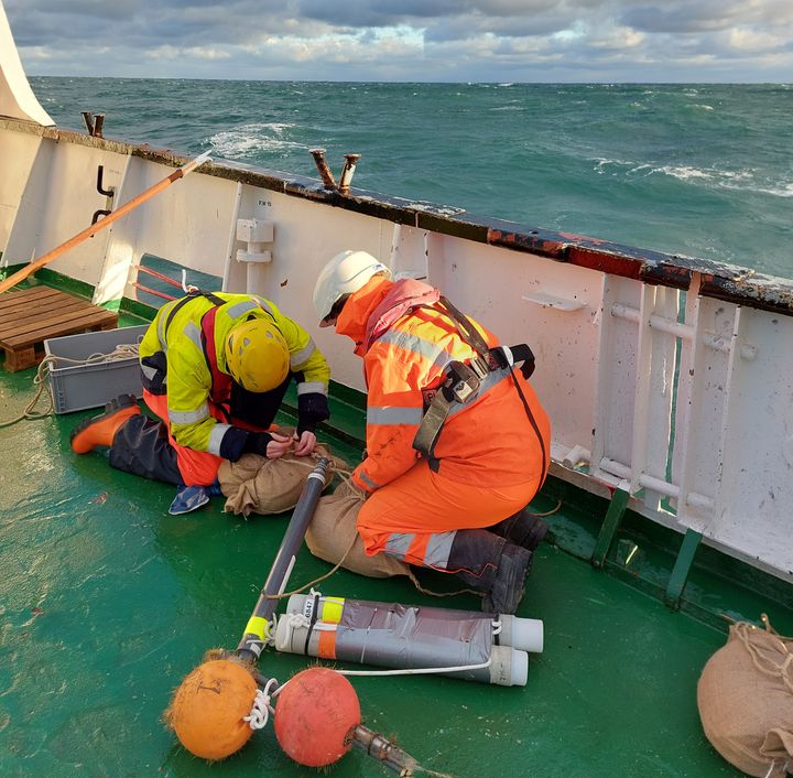 To forskere installerer udstyr på dækket af et skib med havet i baggrunden.