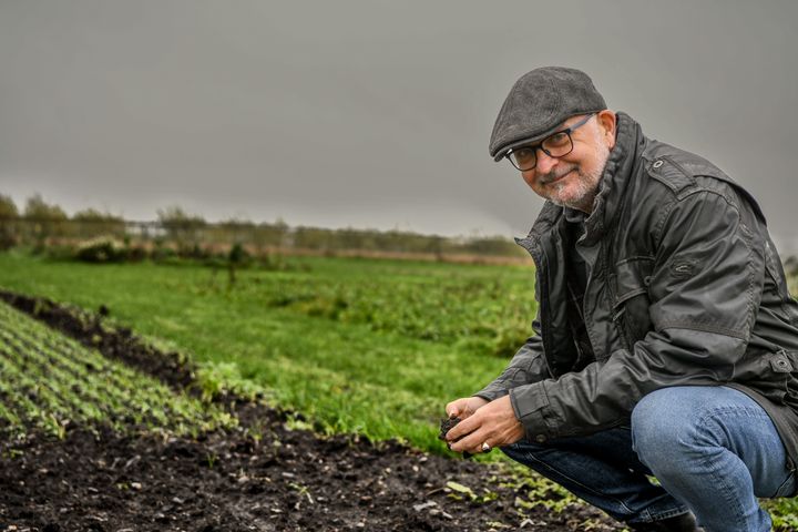 Mogens Greve sidder på hug med hænderne i jorden på en mark, iført hat og jakke, under en overskyet himmel.