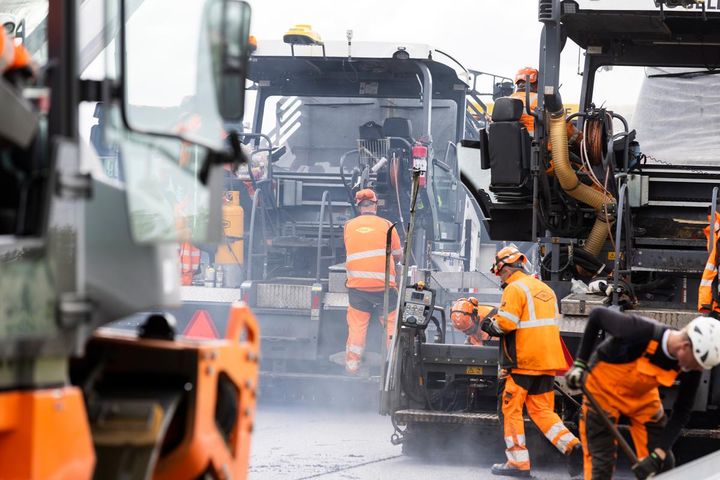 Flere vejarbejdere i orange arbejdstøj arbejder på vejbelægning med store maskiner.