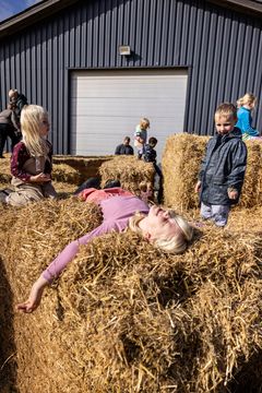 En kvinde og børn leger på halmballer udenfor en lade.