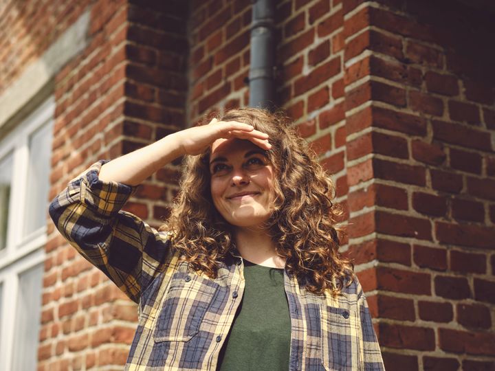 En person står udenfor ved en murstensbygning og skygger for solen med hånden.