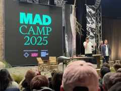 Billede af Gejst projektchef Johan K. Dal, der holder oplæg på en scene med en stor skærm i baggrunden med teksten "MAD CAMP 2025". Publikum ser på.