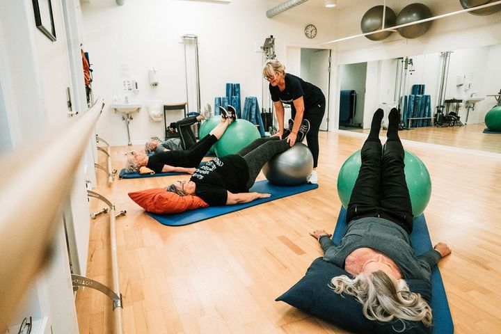 Tre personer udfører øvelser med træningsbolde under vejledning af en instruktør i et træningslokale.