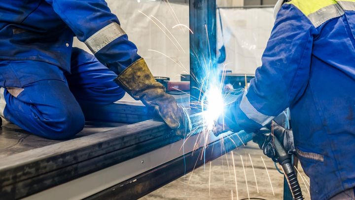 To personer svejser metal iført blå dragter og handsker.