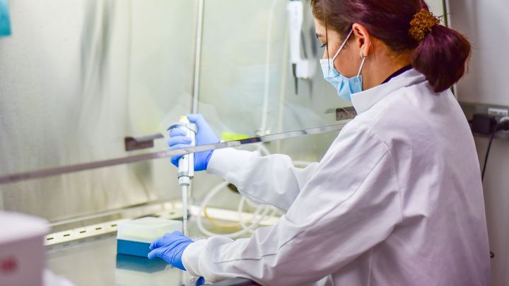 Kvinde af ikke-vestlig oprindelse i kittel og maske arbejder i biolaboratorium med en elektrisk pipette.