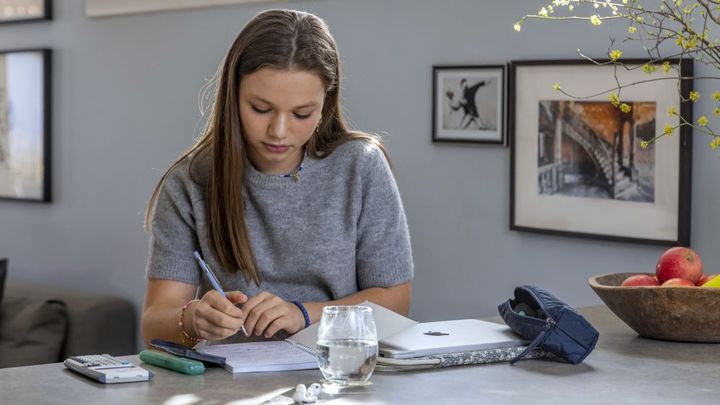 Ung kvinde sidder ved et bord og skriver i en notesbog. En åben bærbar computer og et glas vand står på bordet.