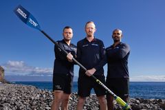 De tre roere står med blå himmel i baggrunden og har hver en hånd på en åre, de holder sammen.