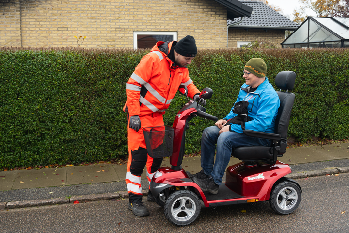 En tekniker fra SOS Dansk Autohjælp i orange tøj hjælper en mand på en rød elscooter.