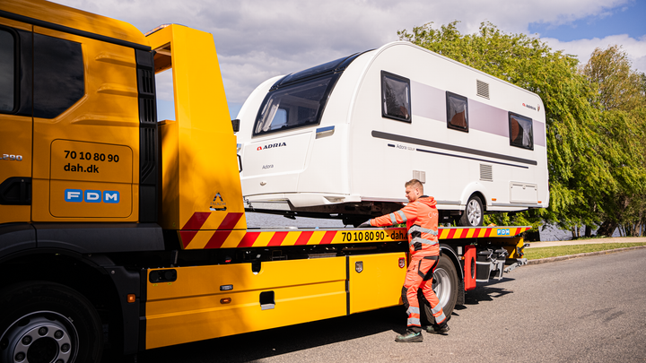 En campingvogn bliver transporteret på en autohjælpevogn fra SOS Dansk Autohjælp på en solrig dag.