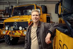 En mand står smilende foran en gul autohjælpsbil i en garage.