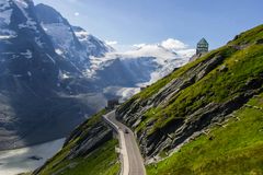 En serpentinväg slingrar sig uppför en grön bergssluttning i ett alpint landskap i Österrike, med snötäckta toppar och en glaciär i bakgrunden.