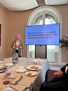 En kvinna står vid ett podie och pratar, med en presentation på en skärm bakom sig.