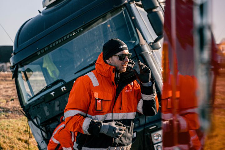 En person i orange arbetskläder står framför en lutande lastbil och talar i en radio.