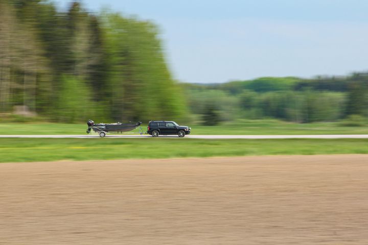En bil trækker en båd langs en landevej med grønne træer i baggrunden.