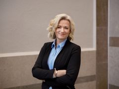 Person i sort blazer og lyseblå skjorte står med armene over kors og smiler.