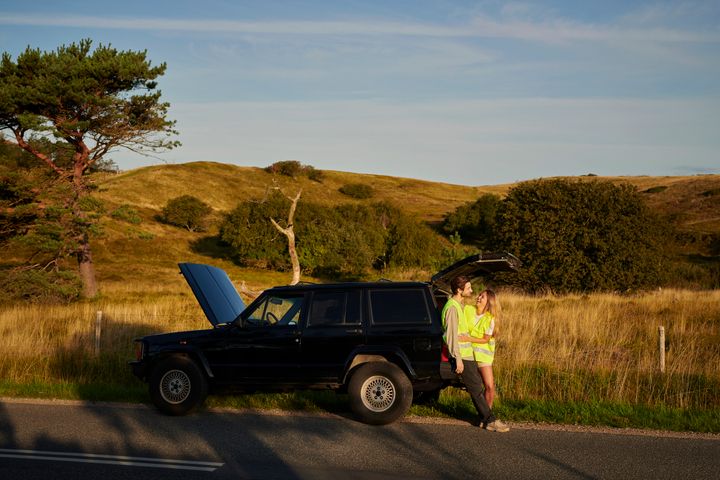Två personer i varselkläder står vid en bil med öppet motorhuv på en landsväg, omgiven av ett landskap med gräs och kullar.